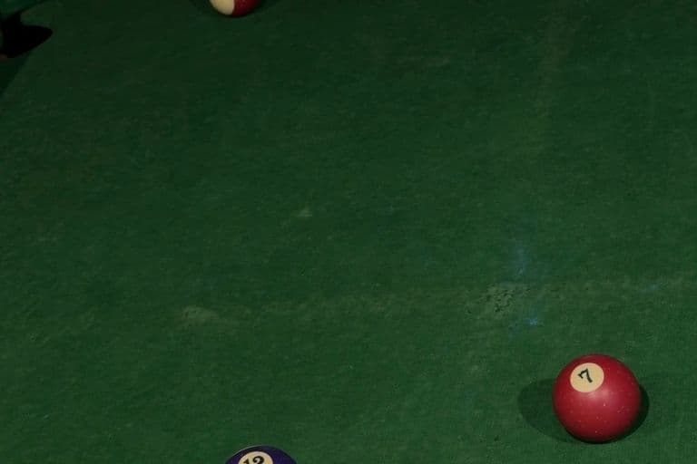 A high-angle, vertical shot of a green felt pool table featuring five billiard balls scattered across the surface. In the foreground, a purple and white striped 12-ball rests on the left, and a solid red 7-ball sits to the right. Further up the table, a maroon and white striped 15-ball is visible, alongside a solid yellow 9-ball near the top left cushion and another solid yellow ball near the top right. The scene is softly lit under dim, overhead lighting.